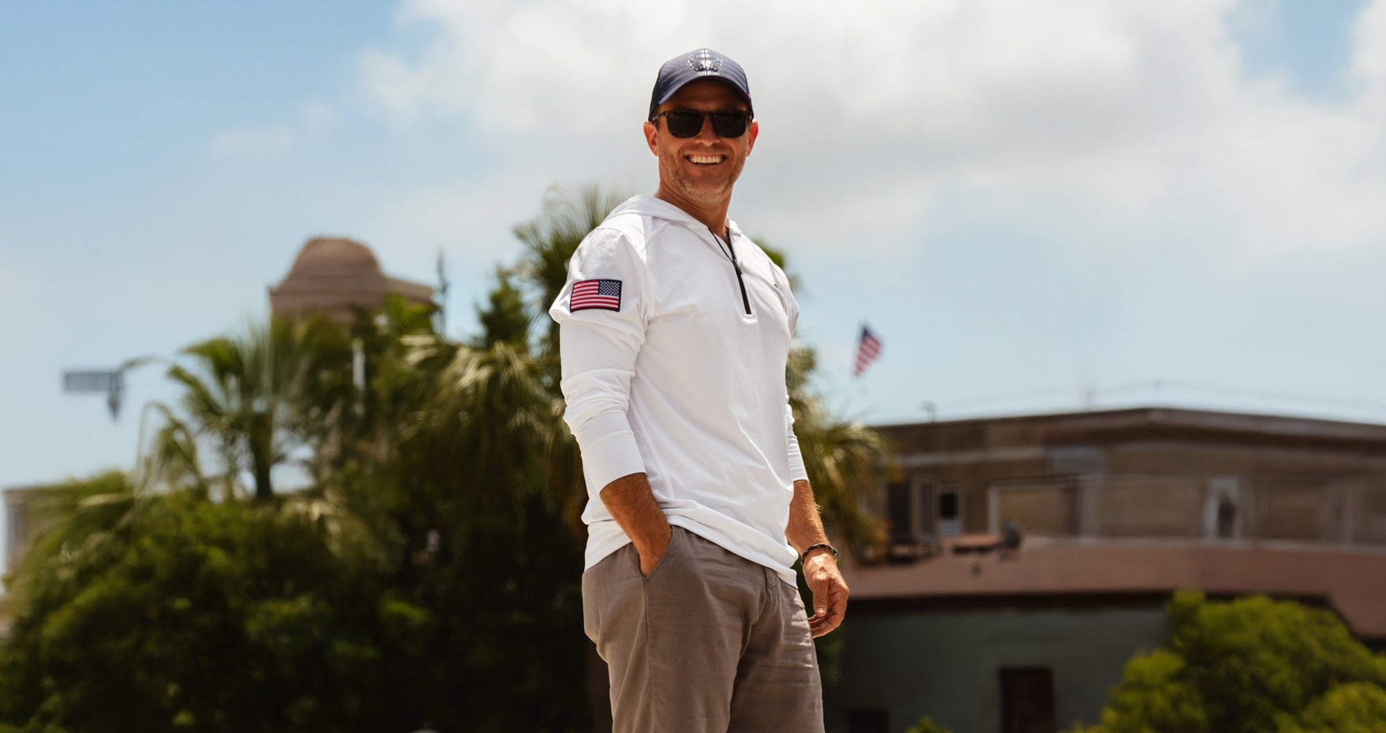 Man in a white tactical hoodie and Volition hat standing outdoors with trees and buildings in the background